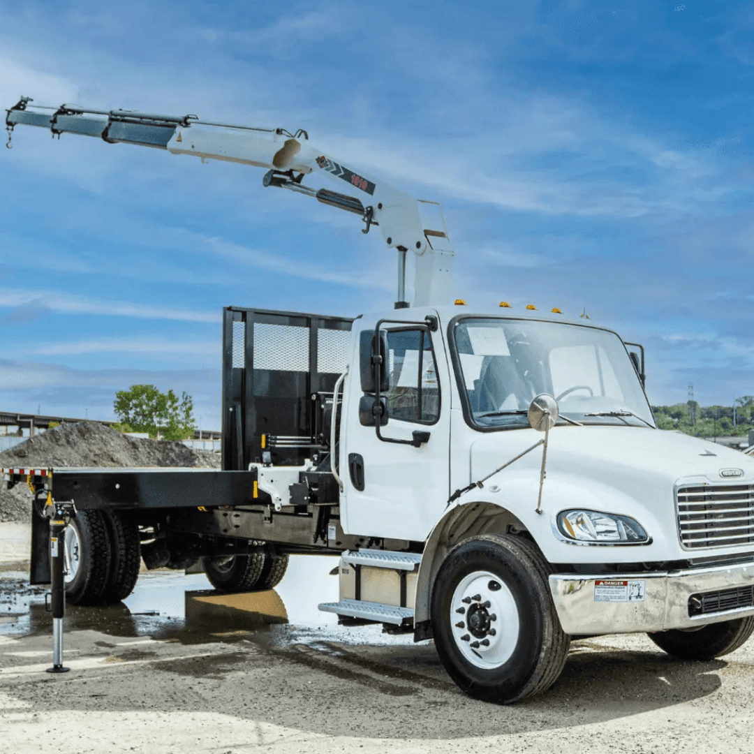 A white boom truck with an extended hydraulic crane arm is parked on a construction site, stabilized by outriggers, with piles of gravel and a blue sky in the background.