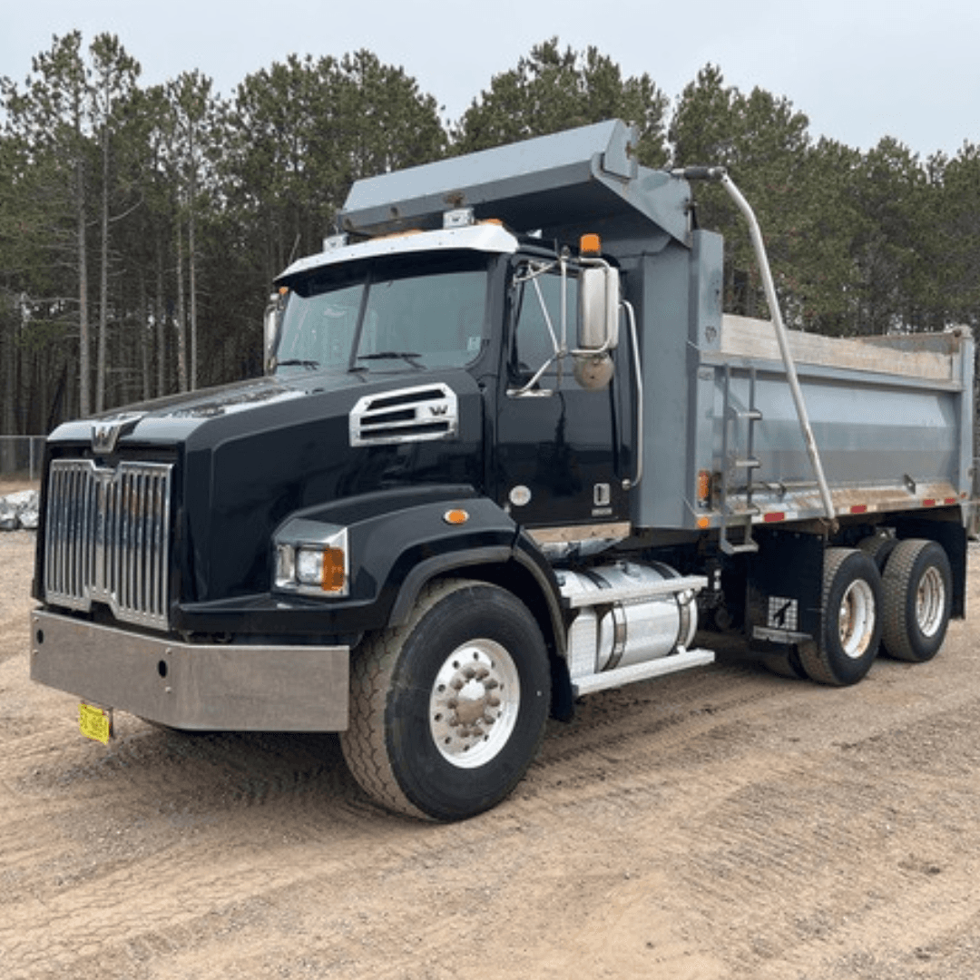 A black and silver dump truck is parked on a dirt lot, with its dump bed raised slightly and trees in the background