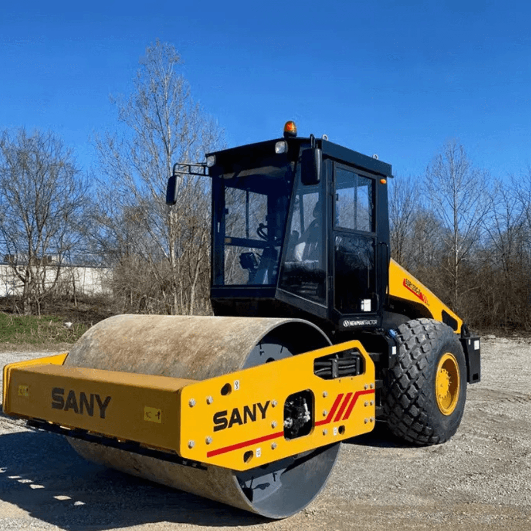 A yellow and black SANY combination roller with a large steel front drum and rear rubber tires sits on a gravel surface, with leafless trees and a clear blue sky in the background