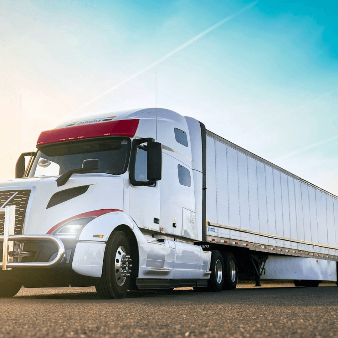 A white semi‑truck with a long enclosed trailer is parked on a paved surface under a bright blue sky.