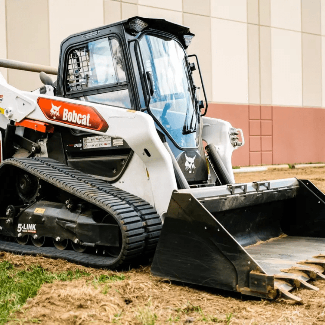 A white and black Bobcat compact track loader with a front bucket attachment sits on a dirt surface beside a commercial building.