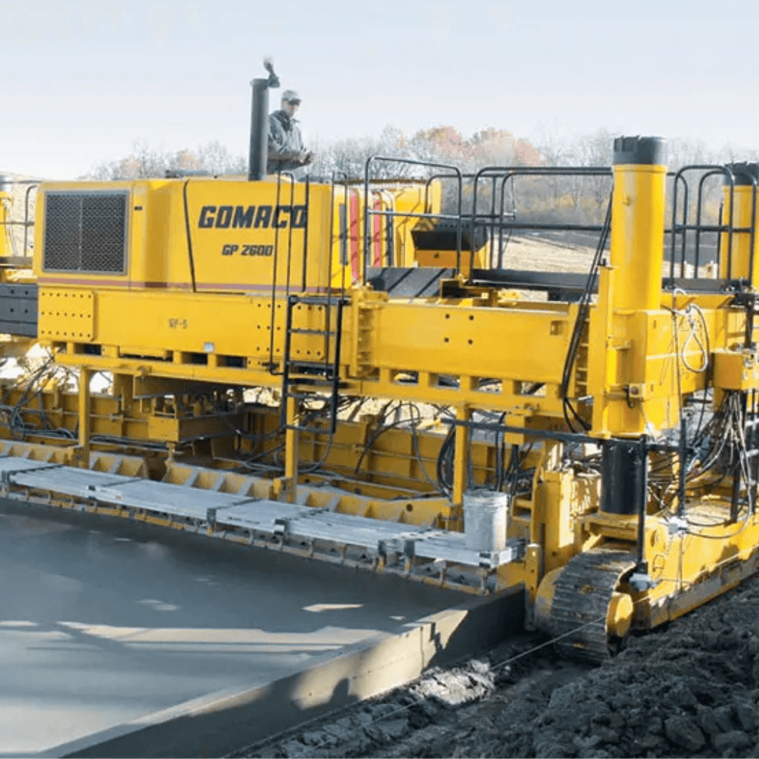 A large yellow GOMACO GP 2600 concrete paver spreads and smooths fresh concrete on a construction site, with an operator standing on the machine and trees in the background
