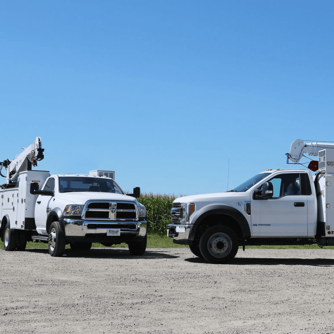 An image of two white utility trucks parked in a dirt lot next to a field under a sunny cloudless sky