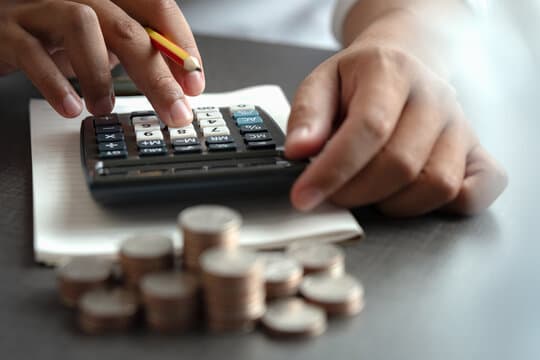 an image with hands holding a pencil typing numbers into a calculator sitting on top of a notepad with coins in front of the calculator