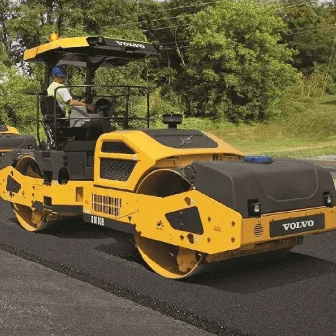 A yellow Volvo asphalt compactor with dual steel drums rolls over freshly laid pavement, operated by a worker wearing a hard hat and safety vest, with trees in the background.