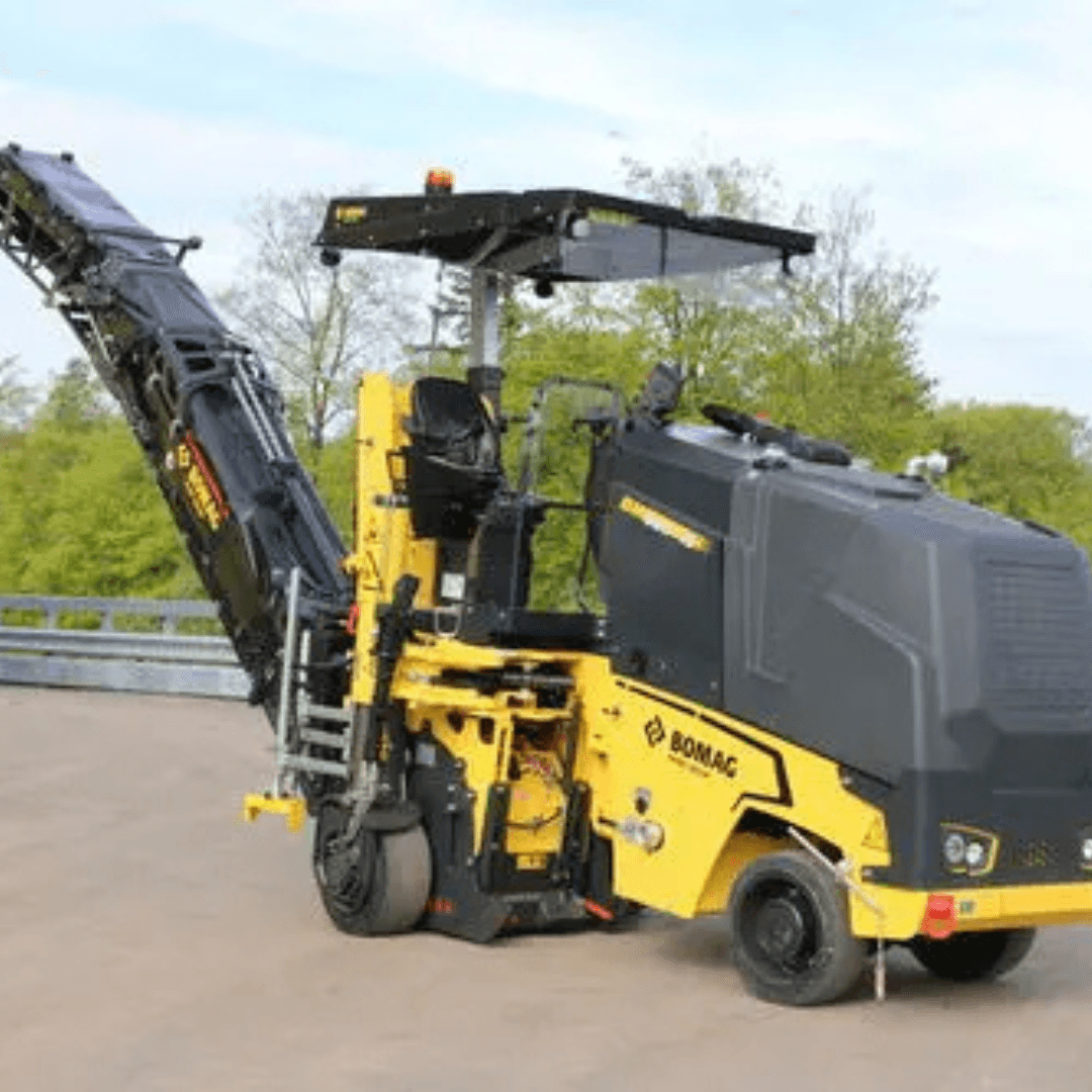 A yellow and black BOMAG cold planer with a long conveyor belt attachment is positioned on a paved surface, with trees in the background under a light sky