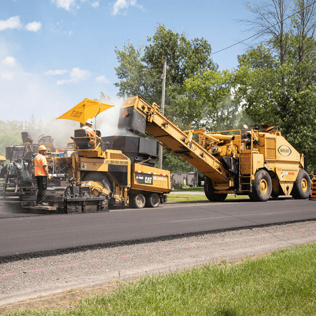 An image of a road paving crew in bright vests running a weiler remixing transfer vehicle and laying new asphalt for a road