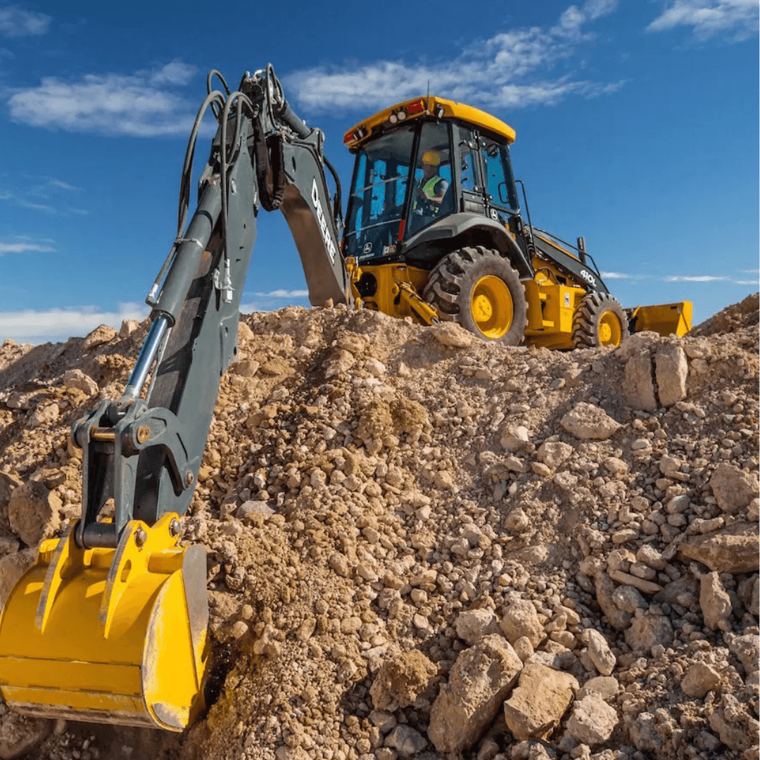 A yellow backhoe loader works on a mound of loose dirt and rocks, with its rear excavator arm extended forward under a bright blue sky