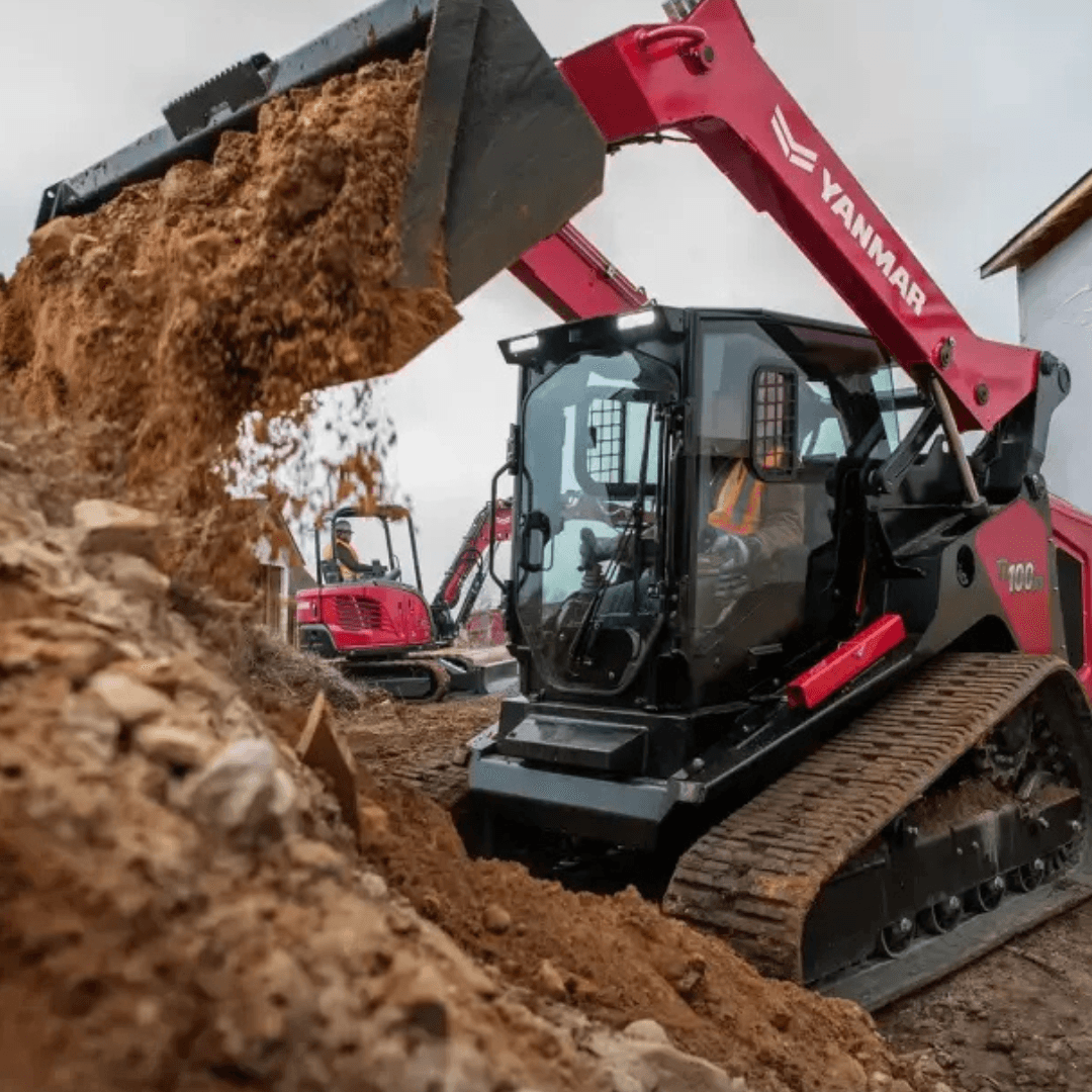 A red and black Yanmar skid steer loader dumps a bucket of dirt on a construction site, with loose soil and another compact machine visible in the background
