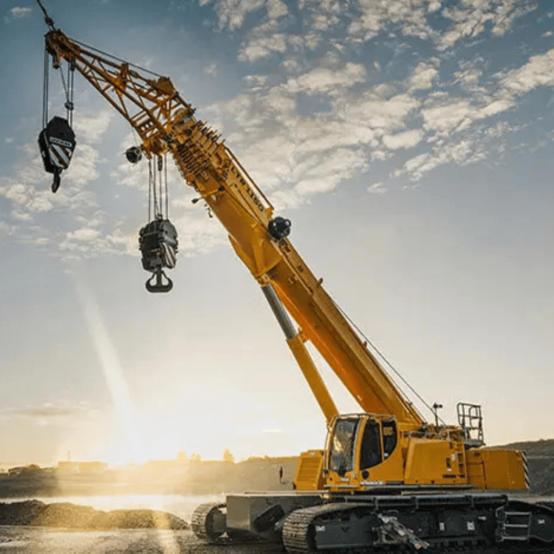 A large yellow crawler crane with an extended telescopic boom sits on tracked undercarriage, lifting heavy hooks at a construction site during sunrise under a partly cloudy sky.