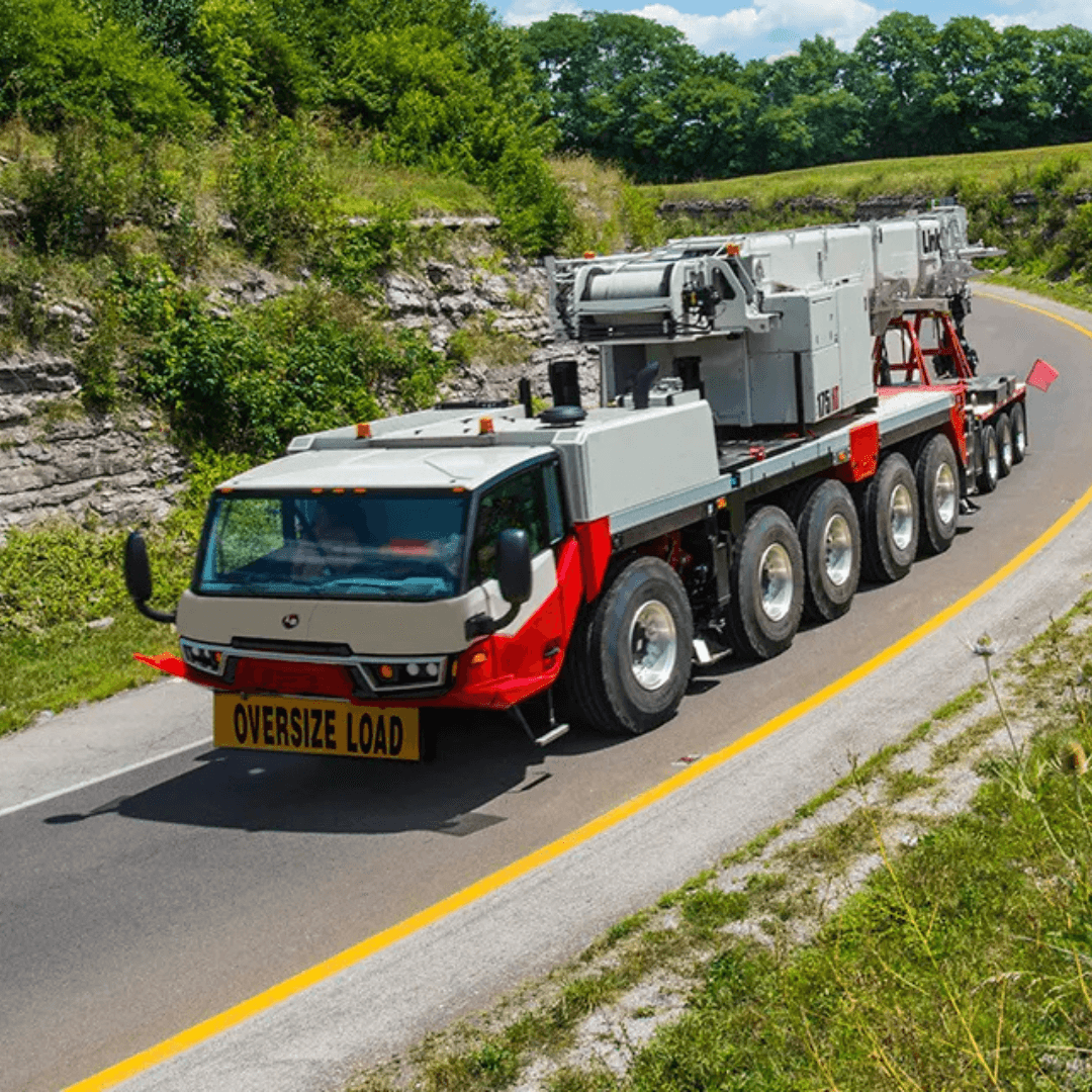 A large multi‑axle all‑terrain crane with an ‘OVERSIZE LOAD’ sign travels down a curved highway, carrying its telescopic boom on the vehicle’s deck, surrounded by rocky embankments and greenery.