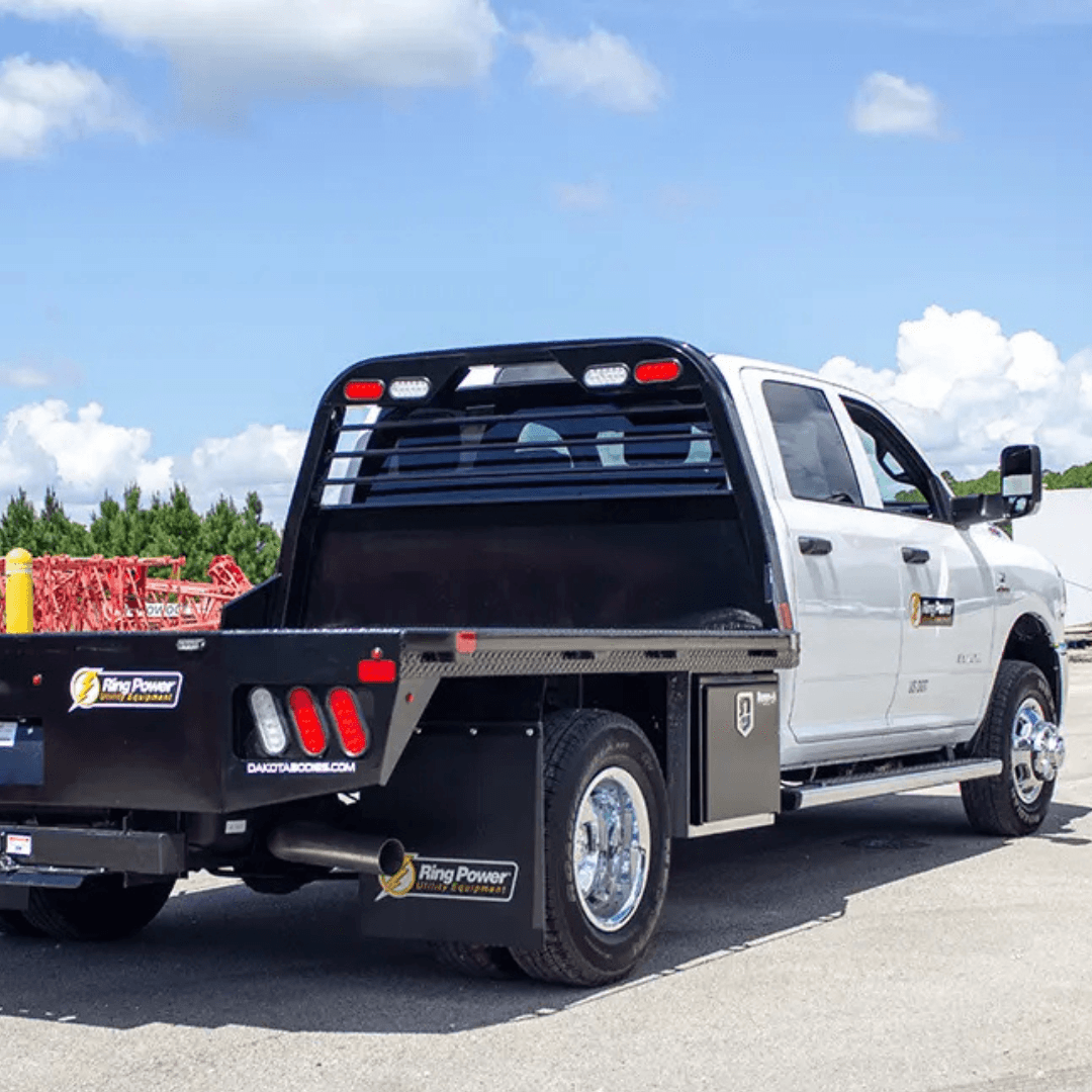 A white work truck with a black flatbed and storage compartments is parked on a paved lot under a partly cloudy sky